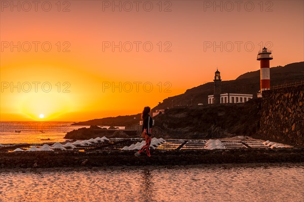 A young woman walking in the orange sunset in the salina and in the background the Fuencaliente Lighthouse on the route of the volcanoes south of the island of La Palma