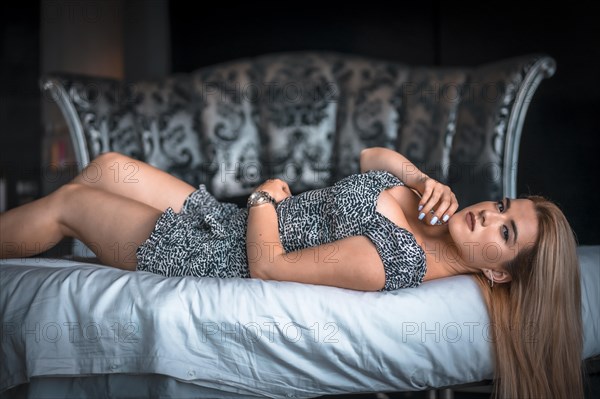 Lifestyle with a young blonde Caucasian woman in a short black and white dress on top of a circular bed with a beautiful headboard. Lying on your back staring intently