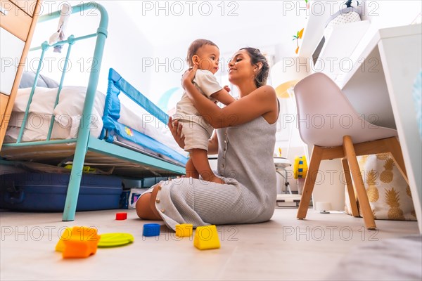 Young Caucasian mother playing with her in the room with toys. Baby less than a year learning the first lessons of her mother. Mother playing with her son and hugging him lovingly