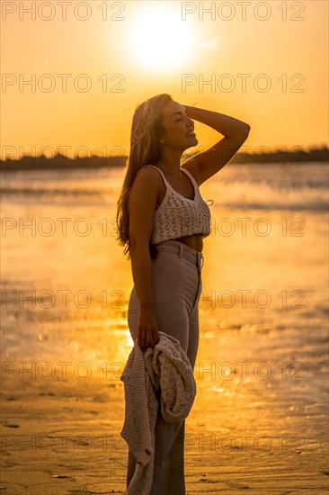 Summer lifestyle. A young blonde Caucasian woman in a white short wool sweater on a beach sunset. With eyes closed and enjoying the sea wind