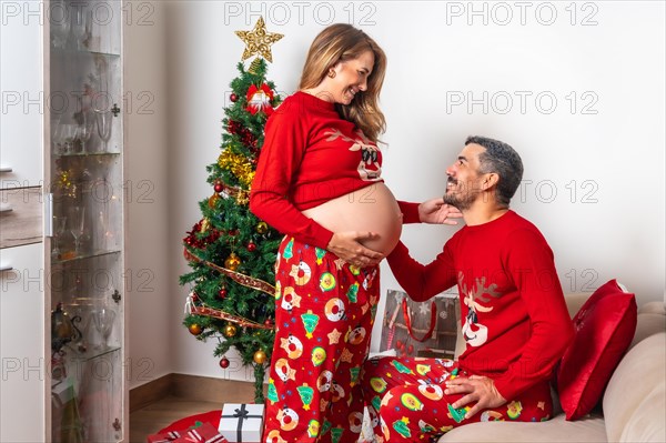 Young couple with decorations and red Christmas clothes looking at each other and very happy for the baby boy who is coming to the family. Family with pregnant woman