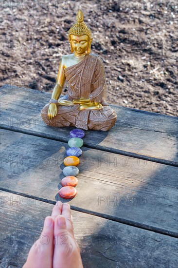 Woman's hands meditating in front of a Buddha statue with colorful chakra stones on a wooden table in a park in the sunshine