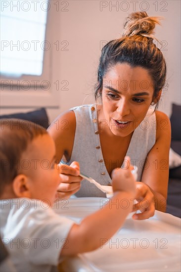 Young Caucasian mother feeding her son a yogurt while sitting in the highchair. Teleworking and caring for your child