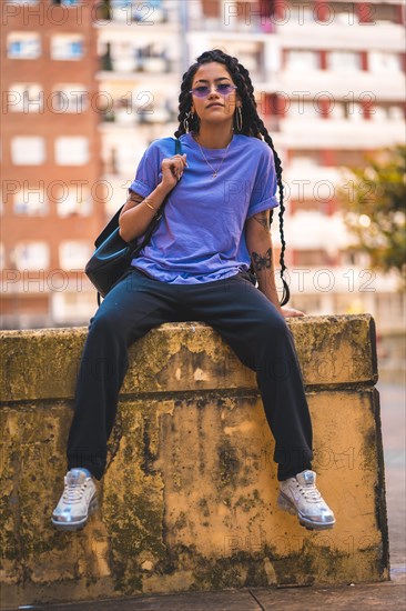 Dark-skinned young woman with long braids and purple glasses sitting on a cement loft