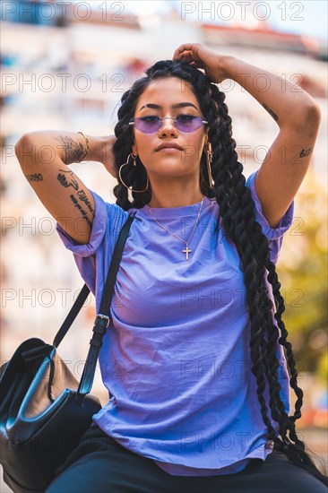 Portrait of young dark-skinned woman with long braids and purple glasses sitting on a bench in the park