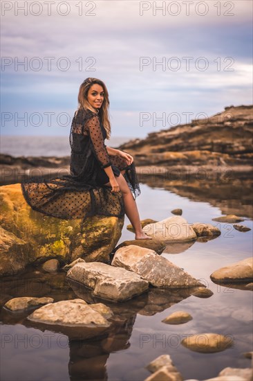 Summer lifestyle with a young brunette Caucasian woman in a long black transparent dress on some rocks near the sea on a summer afternoon. Sitting on top of a rock in a natural pool