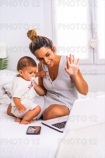 Young Caucasian mother with her son in the room on top of the bed. Drink less than a year in a video call with her family. Communication with grandparents in the confinement of the covid-19 pandemic