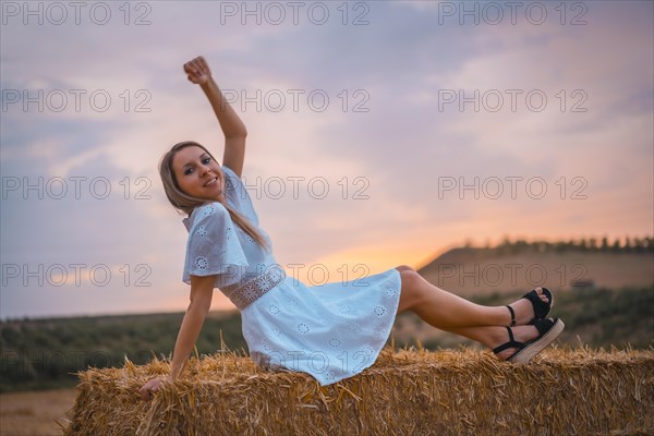 A young blonde Caucasian woman in a white dress in a field of dry straw atop a haystack. In a dry cultivated field in Navarra