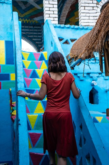 A young woman in a red dress climbing some colorful stairs that go up to a beautiful terrace of a traditional blue house in a Nubian village next to the Nile river and near the city Aswan. Egypt