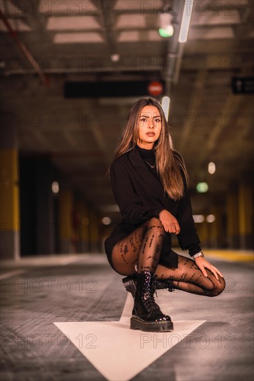 Young caucasian brunette in black jacket on the white arrow of the ground in an empty underground car park. Night urban session in the city