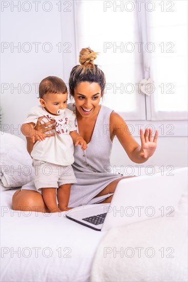 Young Caucasian mother with her son in the room on top of the bed. Drink less than a year in a video call with her family. Communication with grandparents in the confinement of the covid-19 pandemic