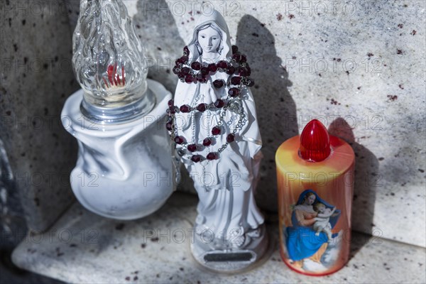 Kitschy electric battery-powered candles next to a small rosary-wrapped figure of Mary on an urn grave in a cemetery