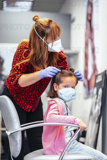 Hairdresser with mask and gloves finishing the pigtails to a girl. Reopening with security measures for hairdressers in the Covid-19 pandemic