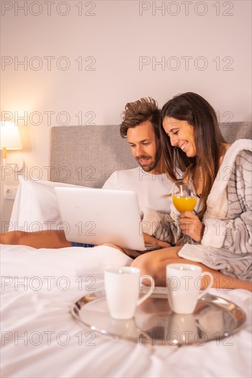 A couple in pajamas looking at the computer in the breakfast of coffee and orange juice in the hotel bed