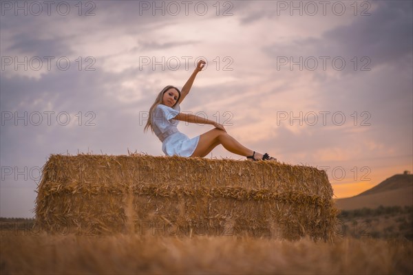 A young blonde Caucasian woman in a white dress in a field of dry straw atop a haystack. In a dry cultivated field in Navarra