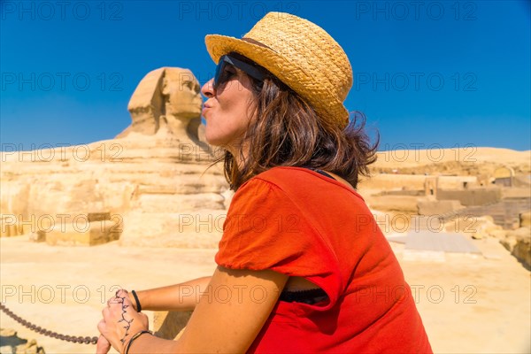 A young tourist in a red dress joking at the Great Sphinx of Giza and in the background the Pyramids of Giza