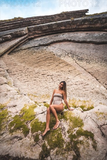 A dark-haired caucasian woman in a brown swimsuit on a natural background next to rocks and sea in the town of Zumaia