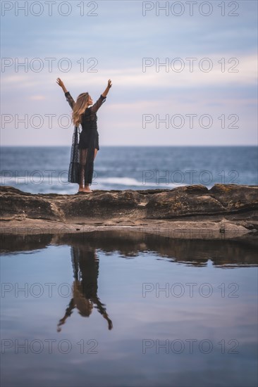 Summer lifestyle with a young brunette Caucasian woman in a long black transparent dress on some rocks near the sea on a summer afternoon. Raising arms and looking out to sea