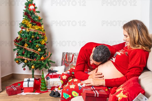 Young couple with decoration and red Christmas clothes listening to the baby's bumps on the belly. Family with pregnant woman