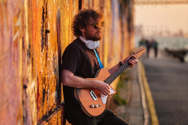 Hipster street musician in black playing electric guitar in the street on sunset leaning on a painted wall