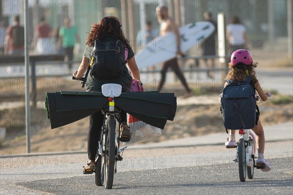 People on the coast: mom and daughter riding bicycles and a surfer with a surfboard. Mid shot