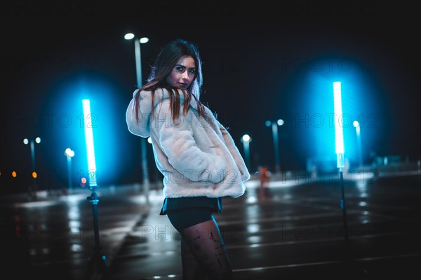 Young Caucasian brunette with pink wool jacket in an empty car park illuminated with blue light. Night urban winter session in the city