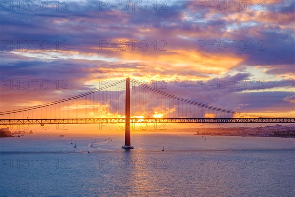 View of 25 de Abril Bridge famous tourist landmark of Lisbon connecting Lisboa and Almada on Setubal Peninsula over Tagus river with tourist yacht boats on sunset. Lisbon