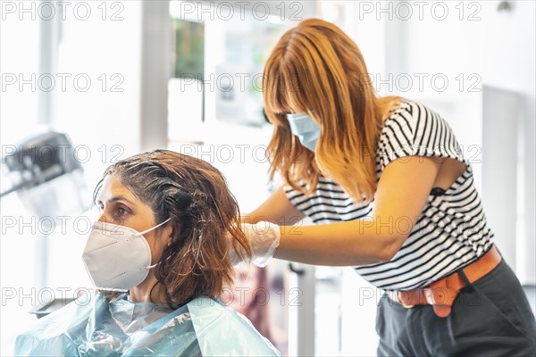 Blonde hairdresser with face mask giving the dark tint to the client at the hairdresser. Safety measures for hairdressers in the Covid-19 pandemic. New normal