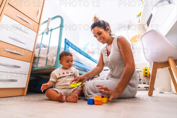 Young Caucasian mother playing with her in the room with toys. Baby less than a year learning the first lessons of her mother. Mother playing with her son playing sitting on the floor