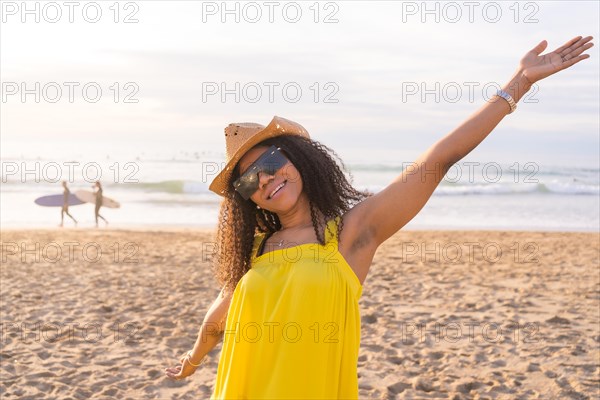 Mixed-raced happy woman wearing sunglasses and sun hat raising arms gesturing well-being on the beach