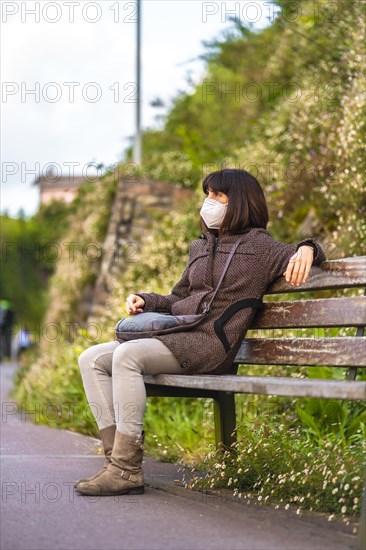 Lifestyle of a young brunette with mask sitting on a wooden bench. First walks of the uncontrolled Covid-19 pandemic