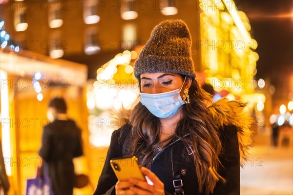 Christmas lifestyle in a new normal. Young girl with face mask visiting the christmas market looking at the mobile phone in the coronavirus pandemic
