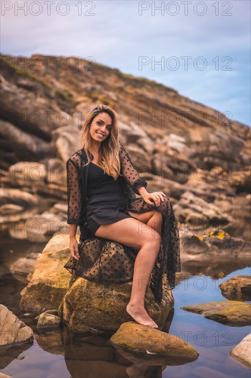 Summer lifestyle with a young brunette Caucasian woman in a long black transparent dress on some rocks near the sea on a summer afternoon. Sitting on a rock smiling and reflected in the water
