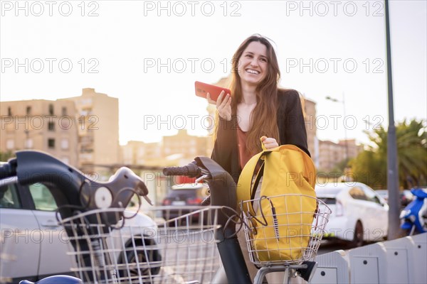 Smiling woman using a rental bike service in the city and talking to the phone