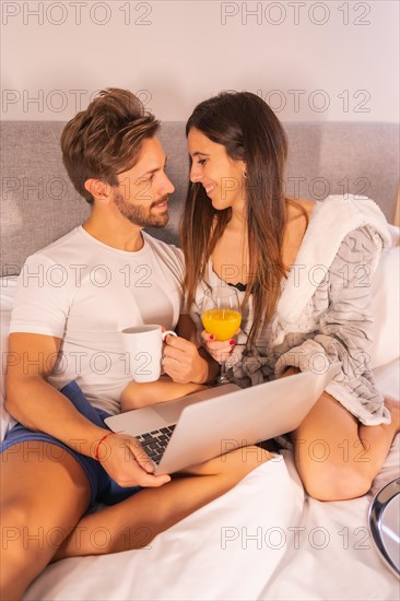 A couple in pajamas looking for hotels or trips from the computer in the breakfast in the hotel bed