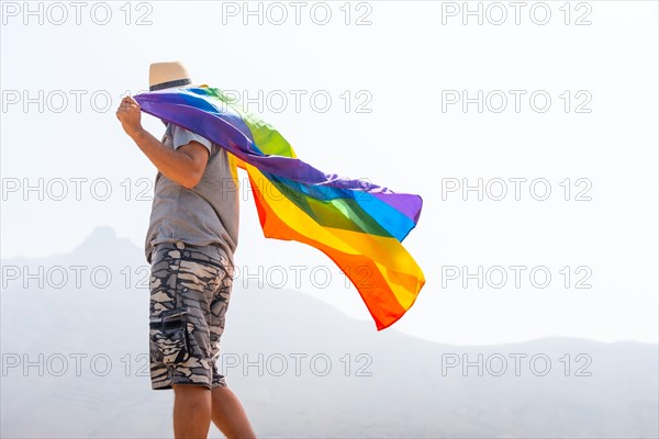 An unrecognizable gay person in a gray t-shirt and white hat waving the LGBT flag in a cloudy sky