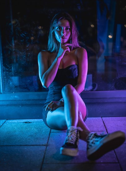 Caucasian brunette in a black dress illuminated by purple and blue led light reflected in black crystals sitting on the floor
