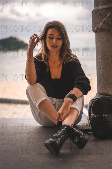Lifestyle in the city with a blonde girl in white pants and a leather jacket near the beach. Sitting on the shallows of a beach looking to the right