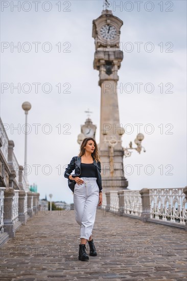 Lifestyle in the city with a blonde girl in white pants and a leather jacket. Perched on a hill with clocks in the background near the beach