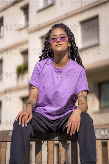 Young dark-skinned woman with long braids in purple glasses sitting on a bench in the park