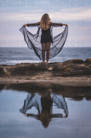 Summer lifestyle with a young brunette Caucasian woman in a long black transparent dress on some rocks near the sea on a summer afternoon. Reflected in the sea