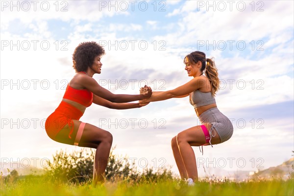 Caucasian blonde girl and dark-skinned girl with afro hair doing squat exercises in a park with the city in the background. Healthy life