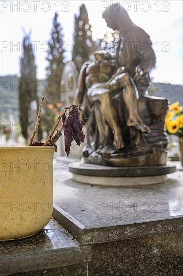 Flowerpot with withered flower on a grave in front of a religious figure of Mary with Jesus in her arms in the backlight
