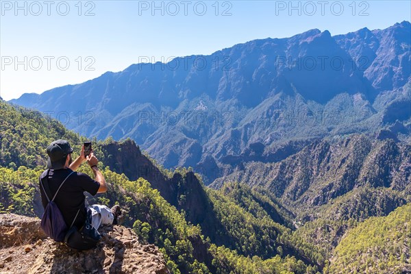 A young man resting after trekking at the top of La Cumbrecita sitting in the natural viewpoint and taking a photo with the mobile