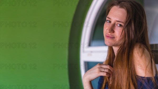 A smiling young pretty redhead Caucasian woman sitting in a blue dress next to a white sale of a green house
