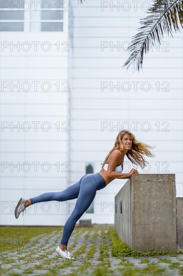 Fitness and yoga session with a young blonde Caucasian instructor dressed in a casual outfit with blue Maya and a white T-shirt. Stretching with a white wall in the background