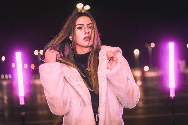 Young Caucasian brunette with wool jacket in an empty car park illuminated with pink light. Night urban winter session in the city