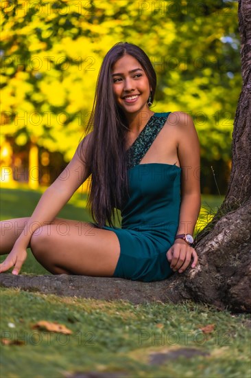 A young pretty brunette Latina with long straight hair leaning against a tree in a green dress. Portrait sitting on the grass next to a tree in the park