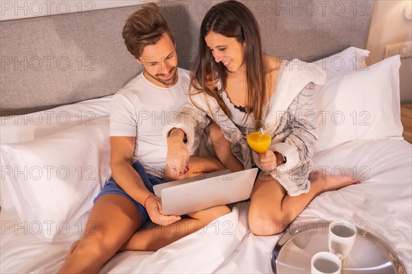 A couple in pajamas looking for hotels or trips from the computer in the breakfast in the hotel bed