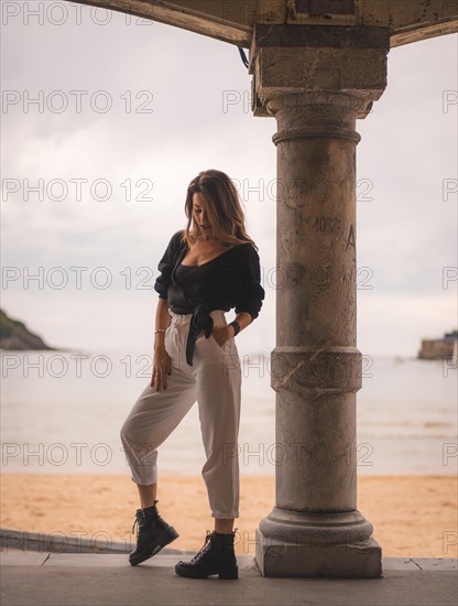 Lifestyle in the city with a blonde girl in white pants and a leather jacket near the beach. Photos next to a column in the shallows of the beach with his arm in his hair